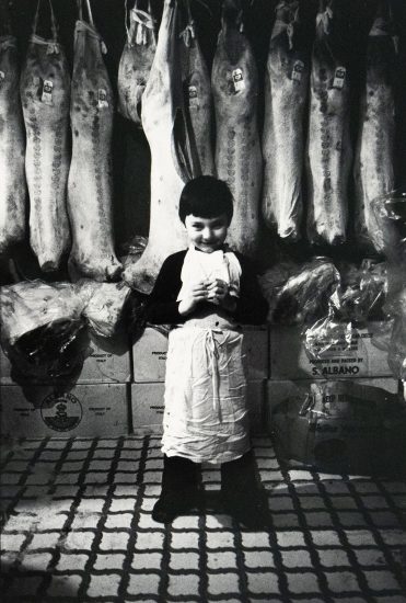 A young boy in a white apron stands smiling in front of hanging sides of meat in a butcher shop. Cardboard boxes labeled "Product of Italy" and "S. Albano" are stacked behind him, and the tiled floor adds to the utilitarian setting.