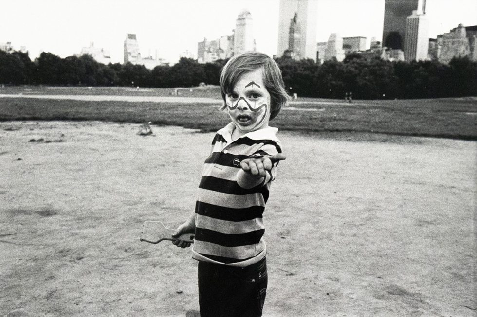 A young child with face paint resembling a clown gestures dramatically toward the camera while holding a paddle, standing on a dirt area in Central Park. The Manhattan skyline rises in the background beyond the tree line.