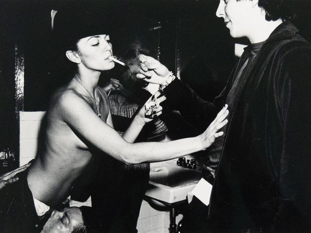 A black and white photograph captures a topless woman in a hat smoking a cigarette, as another person lights it for her. The scene appears to be in a bathroom, with a sink visible in the background.