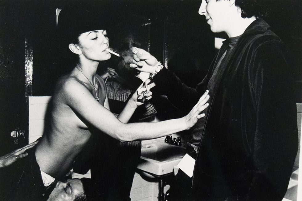 A black and white photograph captures a topless woman in a hat smoking a cigarette, as another person lights it for her. The scene appears to be in a bathroom, with a sink visible in the background.