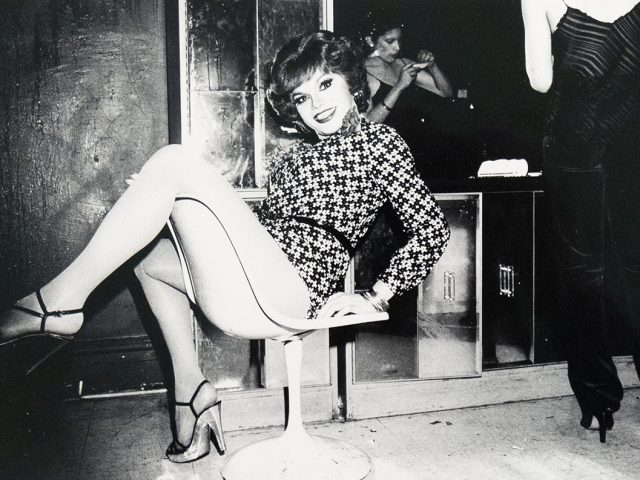 A drag queen with short, wavy hair and a patterned dress sits on a white swivel chair, smiling towards the camera. Her legs are crossed, and she wears strappy heels with hosiery.