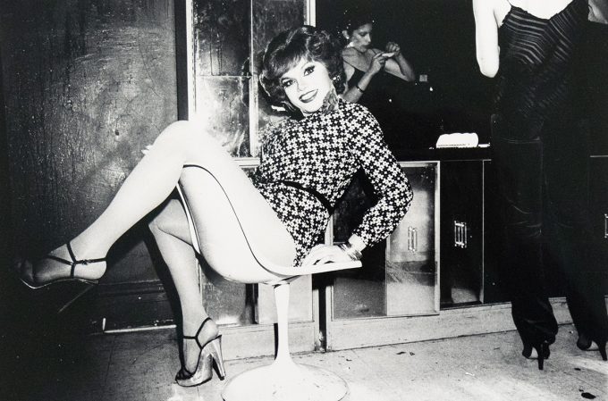 A drag queen with short, wavy hair and a patterned dress sits on a white swivel chair, smiling towards the camera. Her legs are crossed, and she wears strappy heels with hosiery.