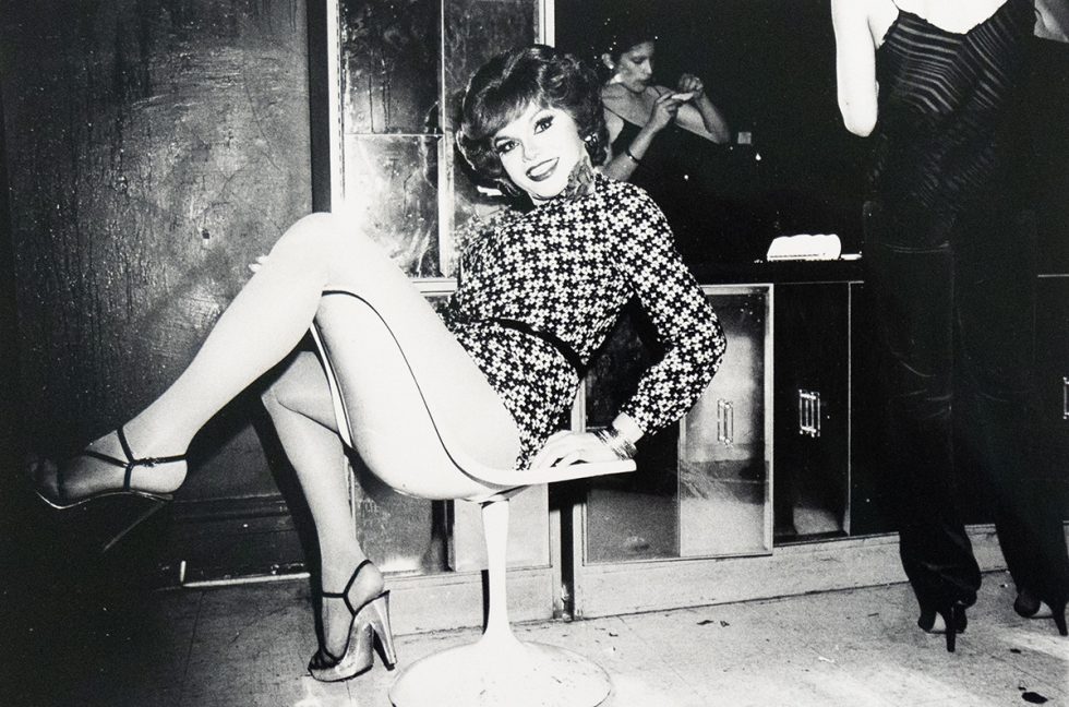 A drag queen with short, wavy hair and a patterned dress sits on a white swivel chair, smiling towards the camera. Her legs are crossed, and she wears strappy heels with hosiery.