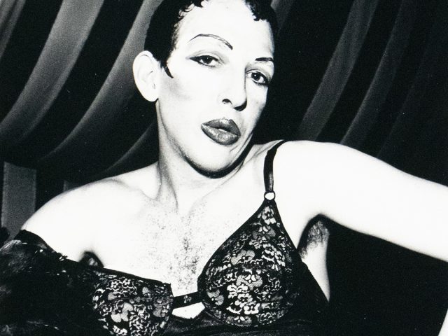 A black-and-white photograph of a drag queen wearing lace lingerie, sitting with legs spread and hands placed on the inner thighs. The drag queen has styled hair, dramatic makeup, and is posed against a background with draped fabric.