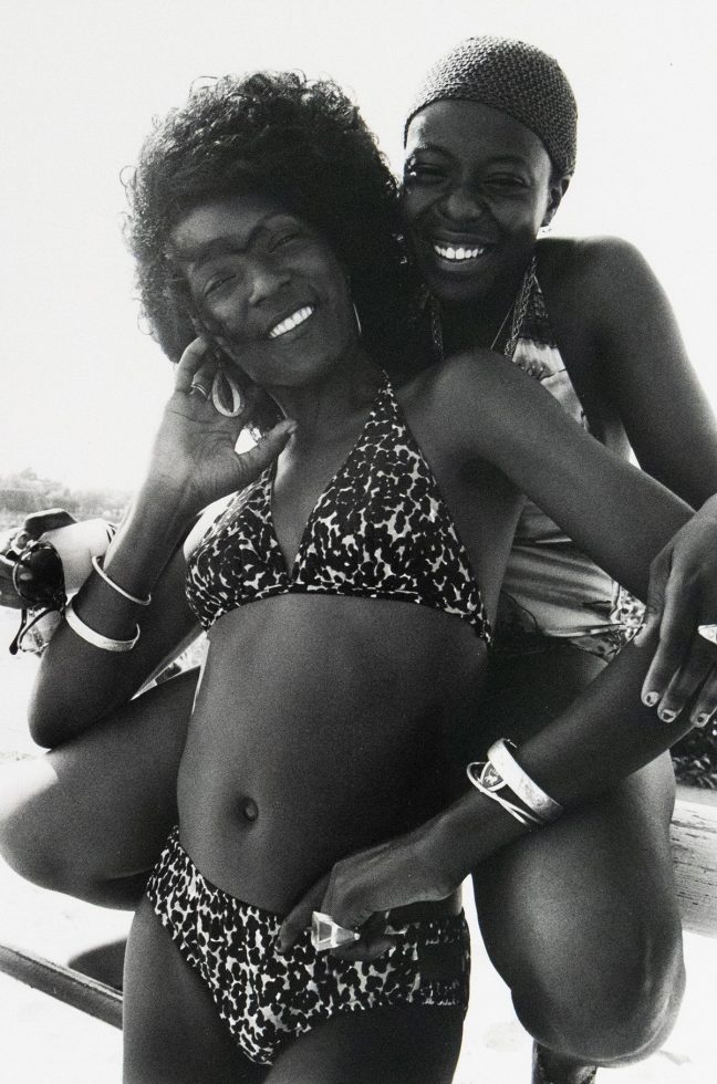 Two women in swimsuits smile closely together, posing outdoors in bright sunlight.