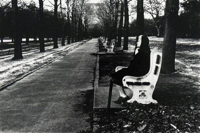 An old woman in dark clothing sits on a white park bench along a paved walkway lined with bare trees. Several empty benches recede into the distance along the walkway.