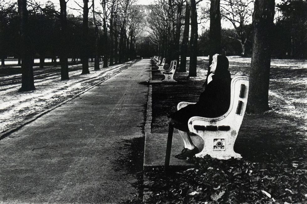 An old woman in dark clothing sits on a white park bench along a paved walkway lined with bare trees. Several empty benches recede into the distance along the walkway.