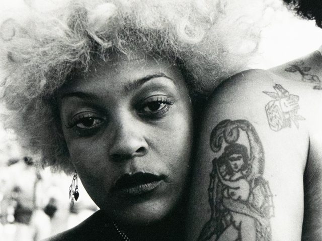 A close-up black and white photograph captures a woman with light, voluminous hair leaning against another person whose arm displays a prominent tattoo of a figure entwined with a snake. The background is bright and softly blurred, suggesting an outdoor setting.
