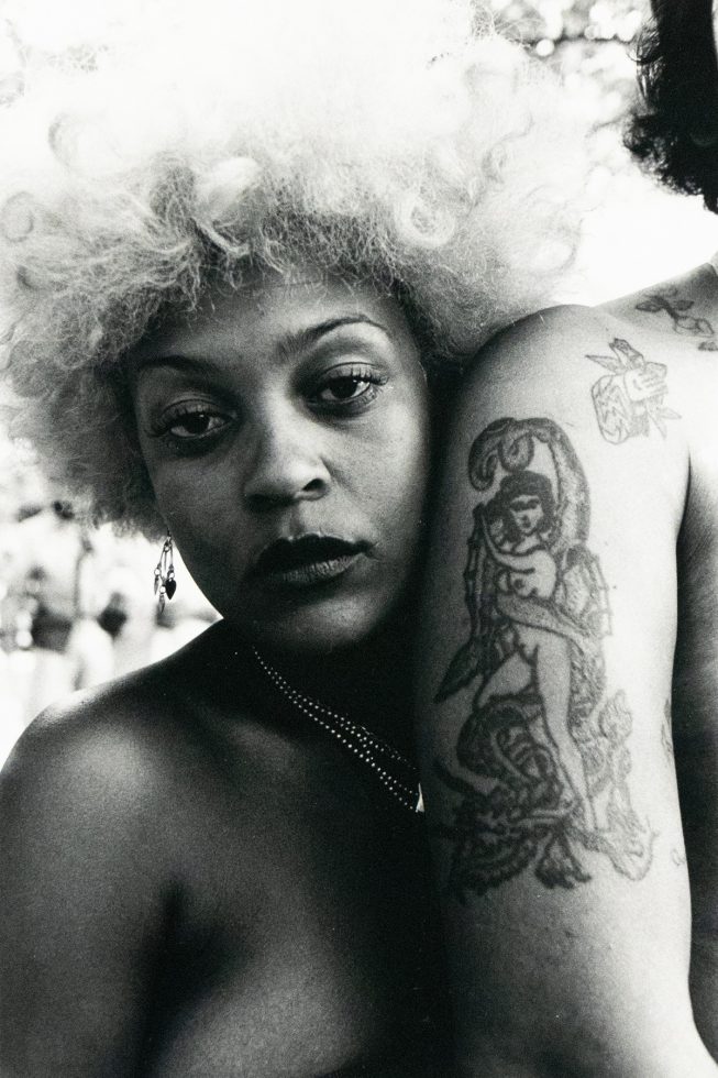 A close-up black and white photograph captures a woman with light, voluminous hair leaning against another person whose arm displays a prominent tattoo of a figure entwined with a snake. The background is bright and softly blurred, suggesting an outdoor setting.