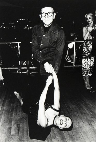 An older man in glasses and a dark jacket smiles while dancing with a woman in a black dress.
