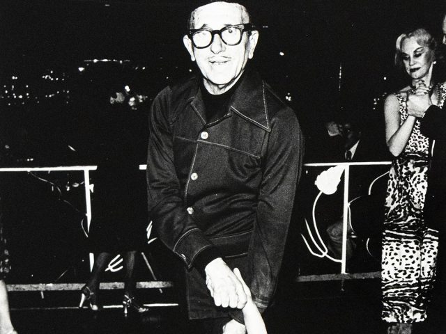 An older man in glasses and a dark jacket smiles while dancing with a woman in a black dress.