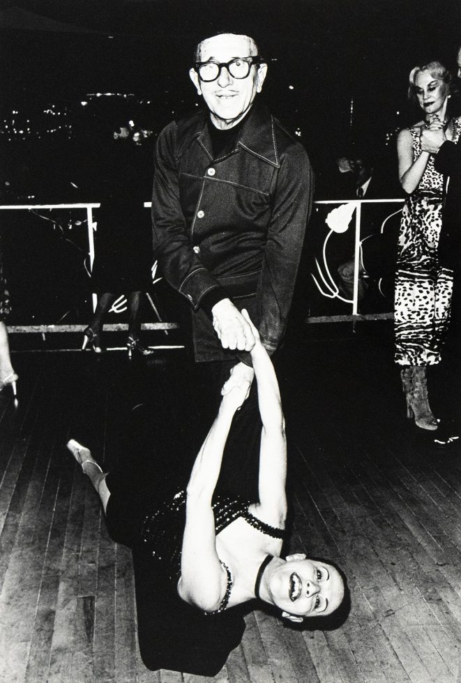 An older man in glasses and a dark jacket smiles while dancing with a woman in a black dress.