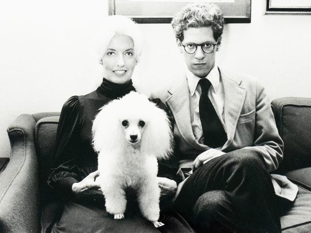 A black-and-white photograph of a woman and a man seated on a couch with a white poodle standing between them. The woman is smiling, and the man is wearing glasses and a suit, looking directly at the camera.