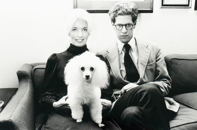 A black-and-white photograph of a woman and a man seated on a couch with a white poodle standing between them. The woman is smiling, and the man is wearing glasses and a suit, looking directly at the camera.