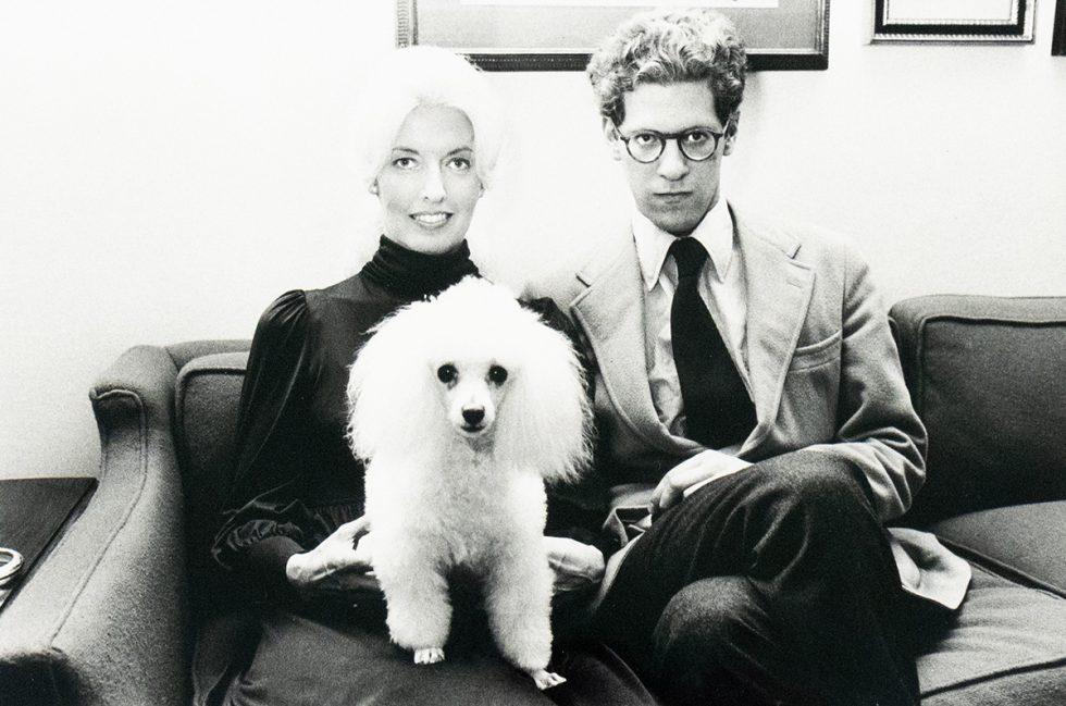 A black-and-white photograph of a woman and a man seated on a couch with a white poodle standing between them. The woman is smiling, and the man is wearing glasses and a suit, looking directly at the camera.