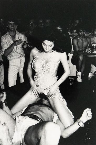 A black and white photograph depicts two figures in an intimate encounter. A man lies on his back, while a woman sits atop his face. The setting is an indistinct, dark indoor space with a crowd of people watching.