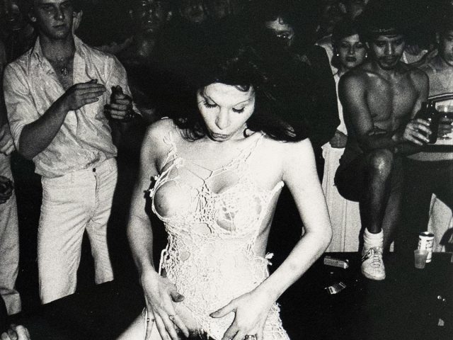 A black and white photograph depicts two figures in an intimate encounter. A man lies on his back, while a woman sits atop his face. The setting is an indistinct, dark indoor space with a crowd of people watching.