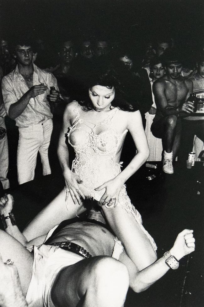 A black and white photograph depicts two figures in an intimate encounter. A man lies on his back, while a woman sits atop his face. The setting is an indistinct, dark indoor space with a crowd of people watching.