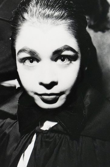 A black-and-white close-up photograph of a woman with slicked-back hair, wide eyes, and a large bow. The lighting creates stark contrasts, emphasizing the woman's expression.