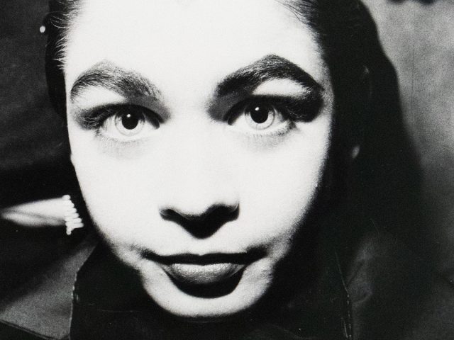 A black-and-white close-up photograph of a woman with slicked-back hair, wide eyes, and a large bow. The lighting creates stark contrasts, emphasizing the woman's expression.
