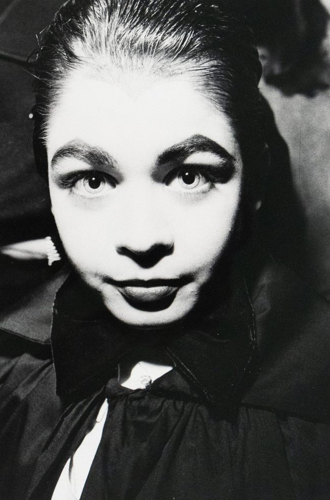 A black-and-white close-up photograph of a woman with slicked-back hair, wide eyes, and a large bow. The lighting creates stark contrasts, emphasizing the woman's expression.