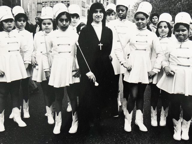 A black-and-white photograph of a group of young girls dressed in matching white marching band uniforms and hats, posing with a nun in a dark habit and large cross necklace. They stand together outdoors on a city street, with buildings and other people in the background.