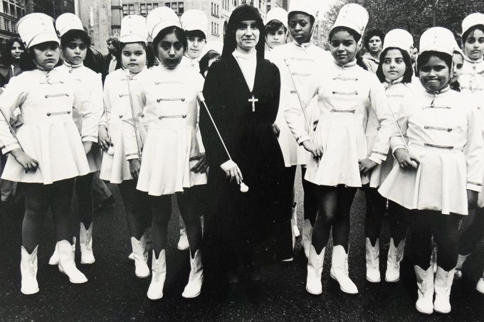 A black-and-white photograph of a group of young girls dressed in matching white marching band uniforms and hats, posing with a nun in a dark habit and large cross necklace. They stand together outdoors on a city street, with buildings and other people in the background.