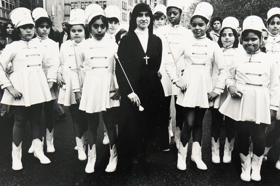 A black-and-white photograph of a group of young girls dressed in matching white marching band uniforms and hats, posing with a nun in a dark habit and large cross necklace. They stand together outdoors on a city street, with buildings and other people in the background.