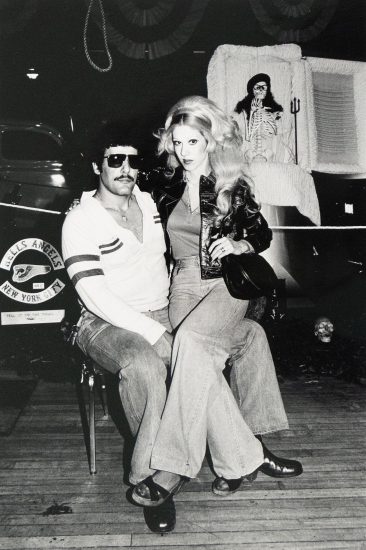 A man with dark hair, a mustache, and sunglasses sits on a chair while a blonde woman in jeans sits on his lap. Behind them is a white coffin with a skeleton figure inside, and a "Hells Angels New York City" sign is visible on a vehicle to the left.