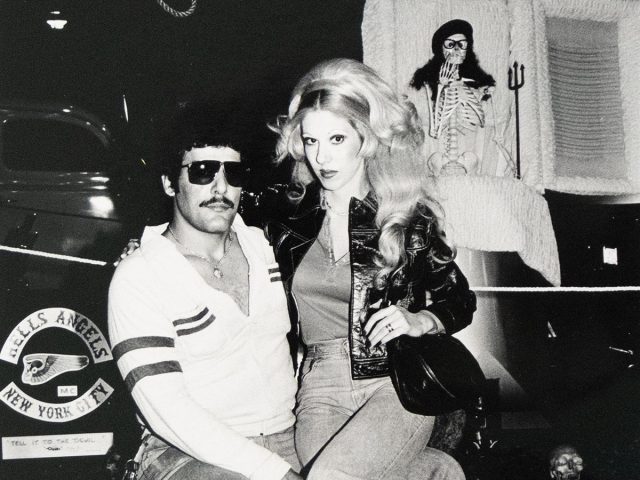 A man with dark hair, a mustache, and sunglasses sits on a chair while a blonde woman in jeans sits on his lap. Behind them is a white coffin with a skeleton figure inside, and a "Hells Angels New York City" sign is visible on a vehicle to the left.