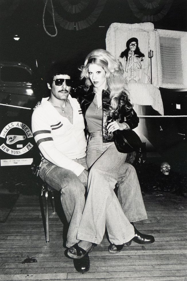 A man with dark hair, a mustache, and sunglasses sits on a chair while a blonde woman in jeans sits on his lap. Behind them is a white coffin with a skeleton figure inside, and a "Hells Angels New York City" sign is visible on a vehicle to the left.