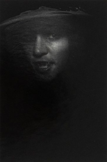 A person's face, partially illuminated, emerges from deep shadow. The subject appears to be wearing a wide-brimmed hat and a vail.