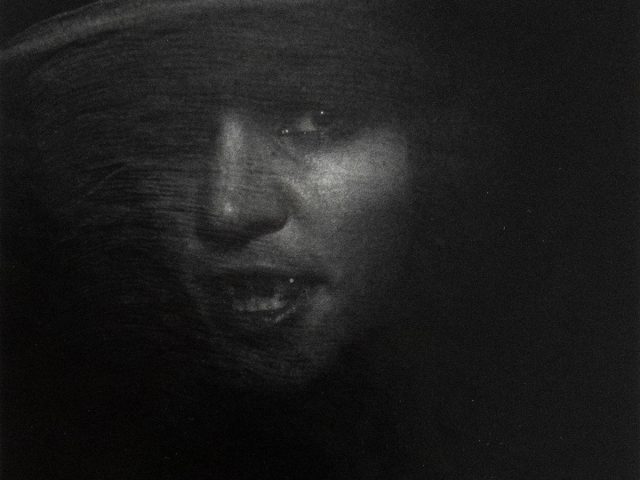 A person's face, partially illuminated, emerges from deep shadow. The subject appears to be wearing a wide-brimmed hat and a vail.