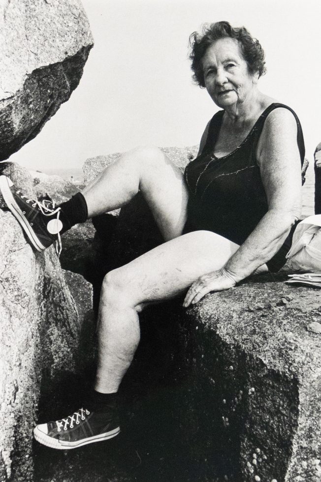 An older woman with short, dark curly hair is sitting on a large rock by the water, wearing a dark swimsuit, black socks, and black and white sneakers