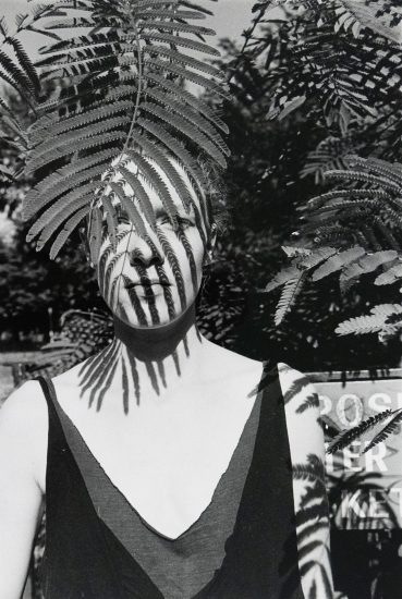 A black-and-white photograph of a woman standing outdoors, partially obscured by fern leaves casting striped shadows across her face and body. A sign with partially visible text appears blurred in the background among foliage.