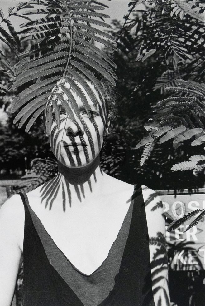 A black-and-white photograph of a woman standing outdoors, partially obscured by fern leaves casting striped shadows across her face and body. A sign with partially visible text appears blurred in the background among foliage.