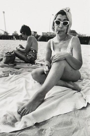 A woman in a swimsuit sits on a beach towel with her legs crossed, wearing cat-eye sunglasses and a towel wrapped around her head, holding a cigarette in her mouth and a lighter in her hand. Another woman sits behind her, turned away, and a boardwalk structure spans the background.