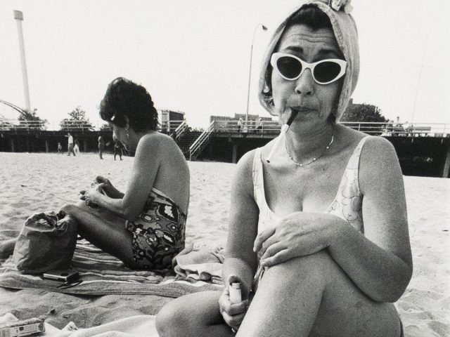 A woman in a swimsuit sits on a beach towel with her legs crossed, wearing cat-eye sunglasses and a towel wrapped around her head, holding a cigarette in her mouth and a lighter in her hand. Another woman sits behind her, turned away, and a boardwalk structure spans the background.