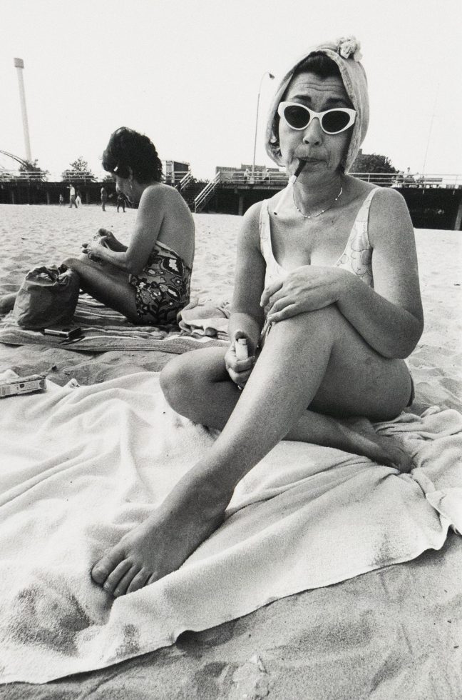 A woman in a swimsuit sits on a beach towel with her legs crossed, wearing cat-eye sunglasses and a towel wrapped around her head, holding a cigarette in her mouth and a lighter in her hand. Another woman sits behind her, turned away, and a boardwalk structure spans the background.