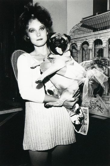 A woman with dark, tousled hair holds a pug dog dressed in a tutu. They are positioned indoors against a backdrop that includes a decorative structure resembling a birdcage or gazebo.