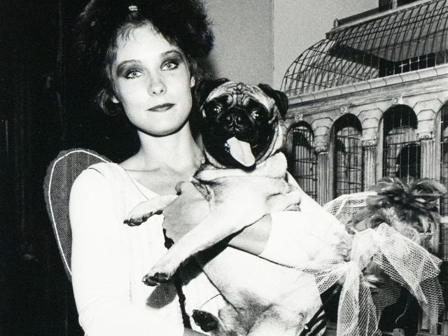 A woman with dark, tousled hair holds a pug dog dressed in a tutu. They are positioned indoors against a backdrop that includes a decorative structure resembling a birdcage or gazebo.