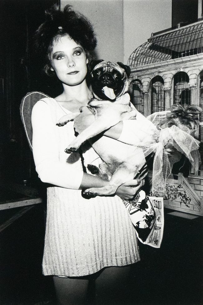 A woman with dark, tousled hair holds a pug dog dressed in a tutu. They are positioned indoors against a backdrop that includes a decorative structure resembling a birdcage or gazebo.