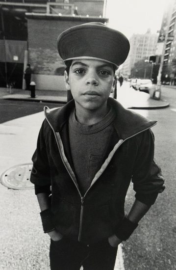 A young person wearing a wide brim hat and half-zipped jacket stands with his hands in his pockets on a city street. Buildings and a crosswalk signal are visible in the blurred background.