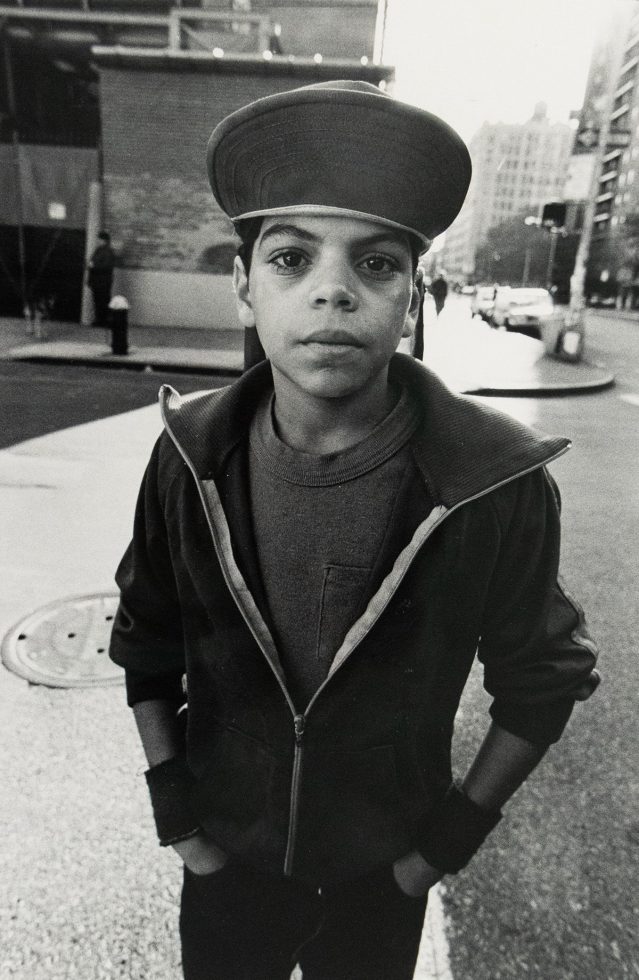 A young person wearing a wide brim hat and half-zipped jacket stands with his hands in his pockets on a city street. Buildings and a crosswalk signal are visible in the blurred background.