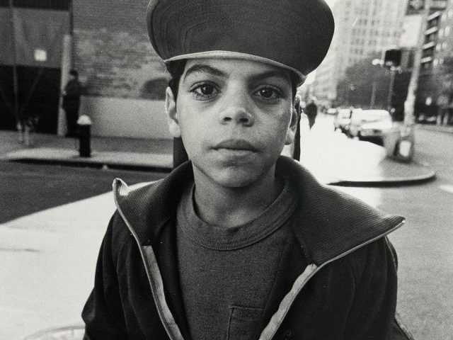 A young person wearing a wide brim hat and half-zipped jacket stands with his hands in his pockets on a city street. Buildings and a crosswalk signal are visible in the blurred background.