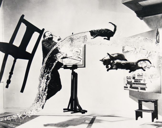 A man with a paintbrush in his mouth leans back as water, cats, and a chair fly across a studio filled with paintings.