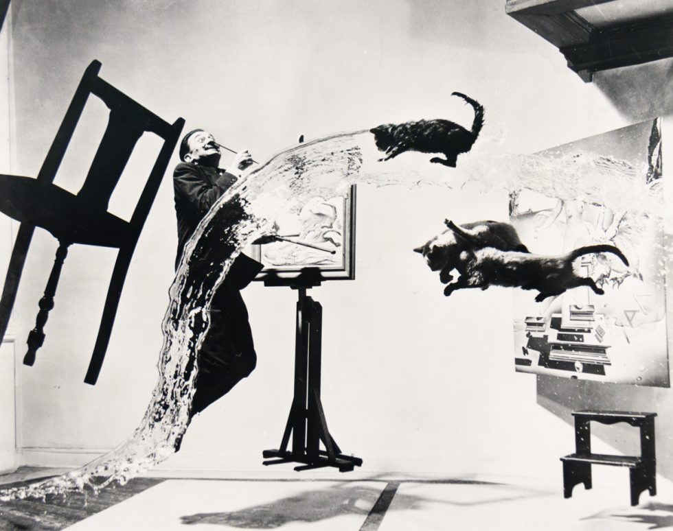 A man with a paintbrush in his mouth leans back as water, cats, and a chair fly across a studio filled with paintings.