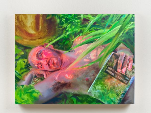 A man lies nude among lush greenery, holding a reflective book partially obscured by leaves.