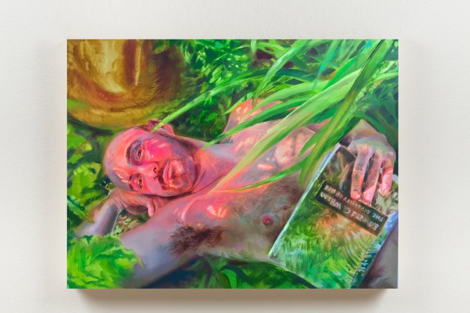 A man lies nude among lush greenery, holding a reflective book partially obscured by leaves.