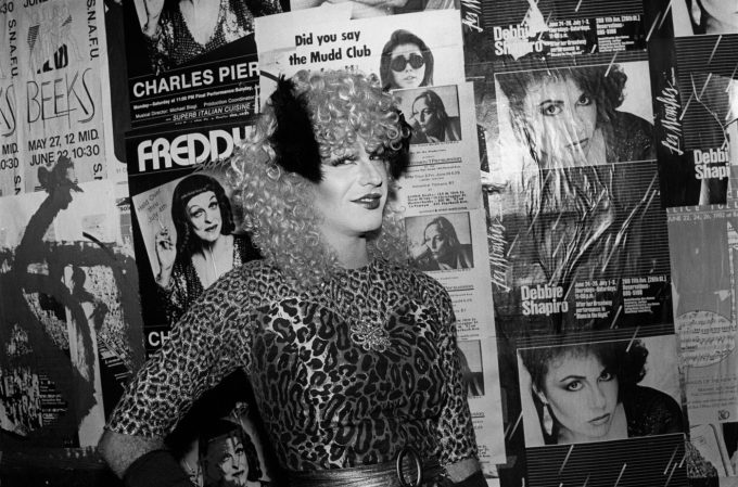 A drag artist with curly blonde hair, a feathered headpiece, and a leopard-print outfit poses in front of a wall covered in concert and club posters.